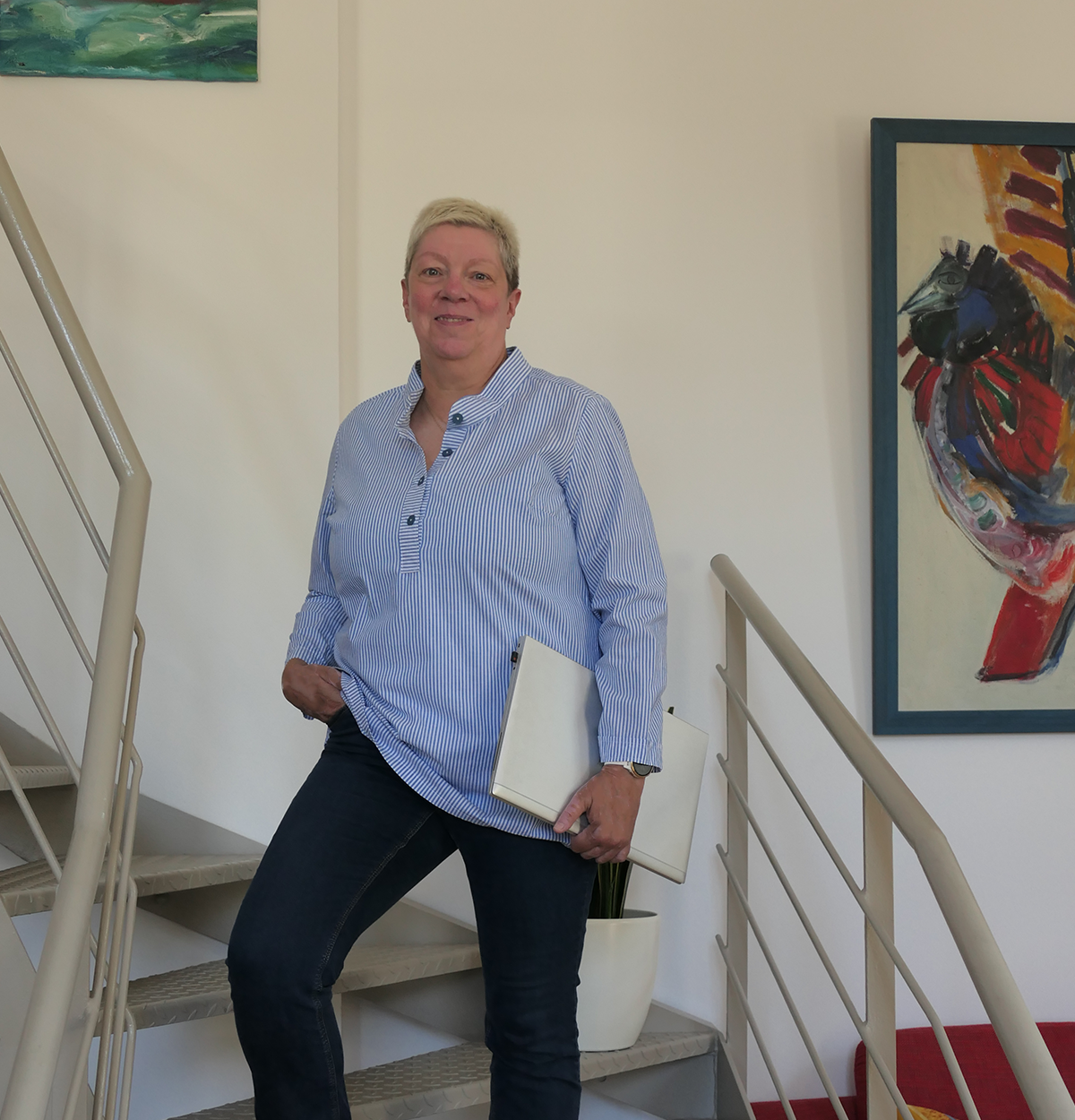 Ute Dreher von Plan D Plan D-Inhaberin Ute Dreher in blau-weiß-gestreiftem Blouson mit einem Laptop in der Hand auf einer Metalltreppe mit Gemälden an der Wand dahinter